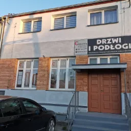 Fasada budynku firmy Cortex oferującej drzwi i podłogi, z brązowymi drzwiami wejściowymi, oknami wystawowymi z ekspozycją produktów i reklamowym banerem nad wejściem, zaparkowany czarny samochód...