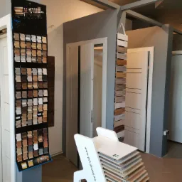 Ekspozycja próbek kolorów i materiałów drzwi oraz podłóg w salonie, z widocznymi skrzydłami drzwiowymi w tle i stojakiem z próbkami paneli podłogowych na pierwszym planie.