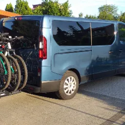 Niebieski van Renault z zamontowanym bagażnikiem rowerowym Thule, przewożący cztery rowery górskie na tle brukowanej posesji z zadaszeniem i ogrodzeniem.