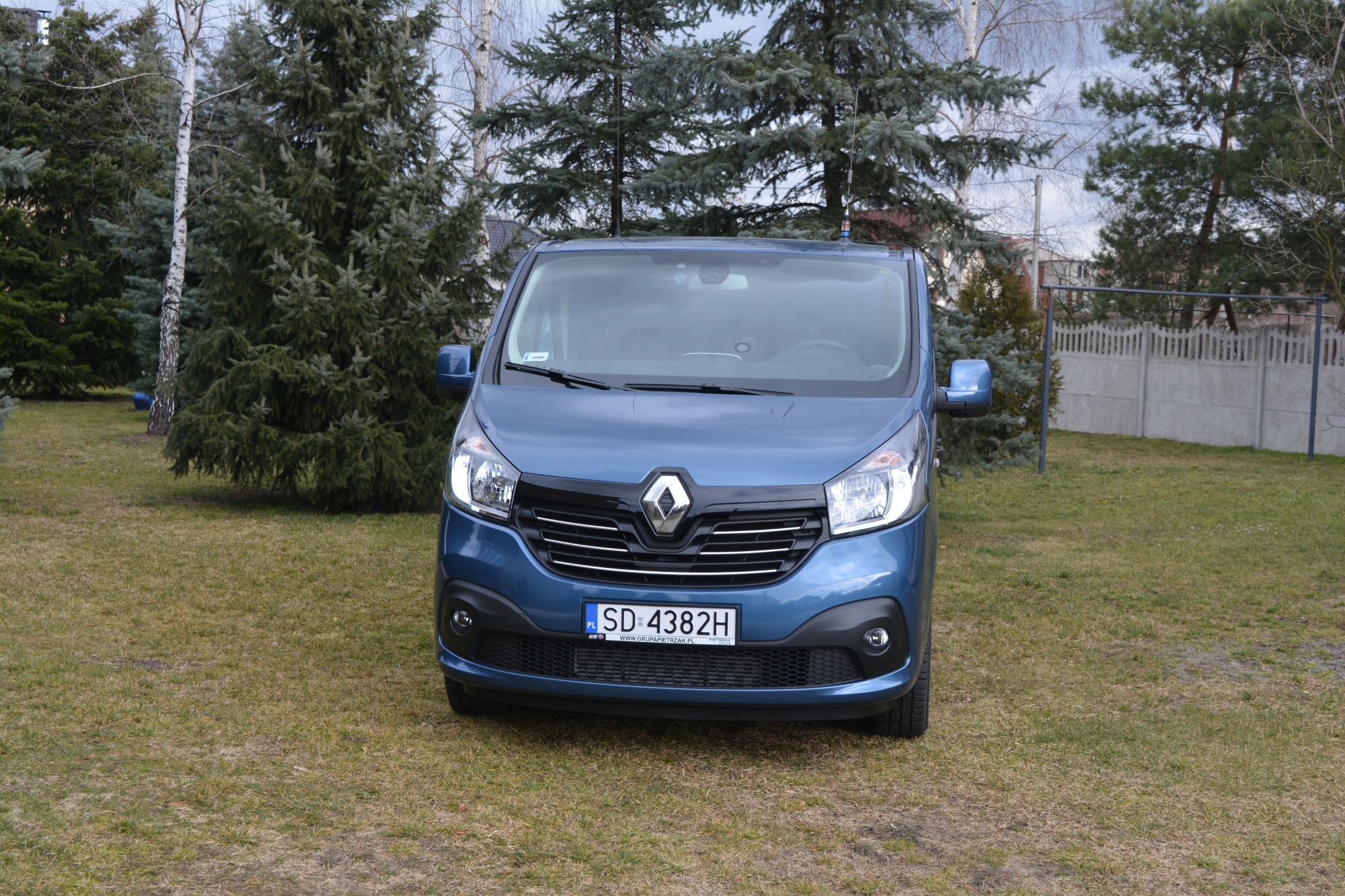 Niebieski bus Renault Trafic zaparkowany na trawniku przed domem, widok z przodu, antena CB na dachu.