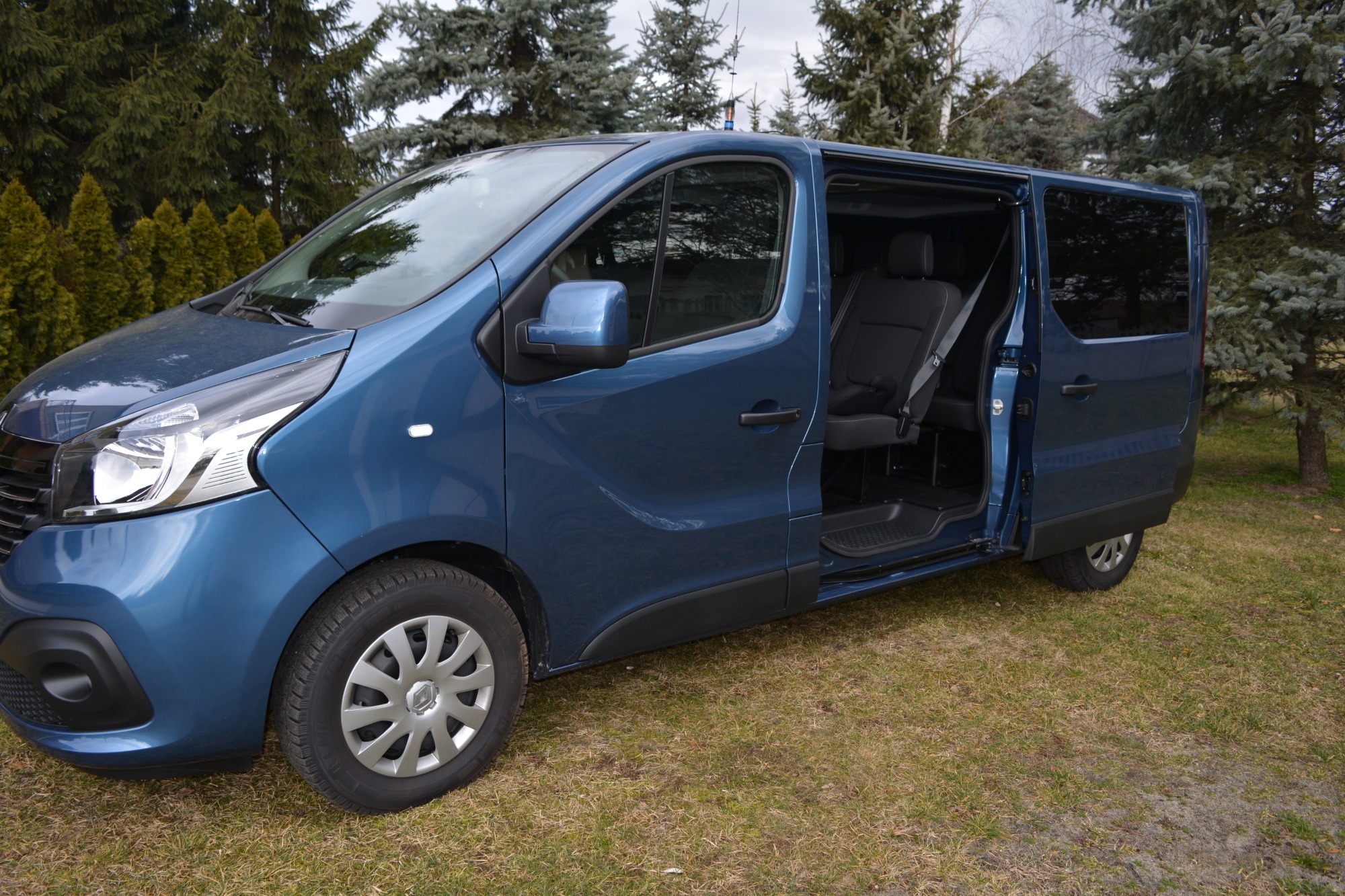 Niebieski van z otwartymi drzwiami przesuwnymi, widoczne fotele pasażerskie, na trawniku w tle drzewa iglaste.