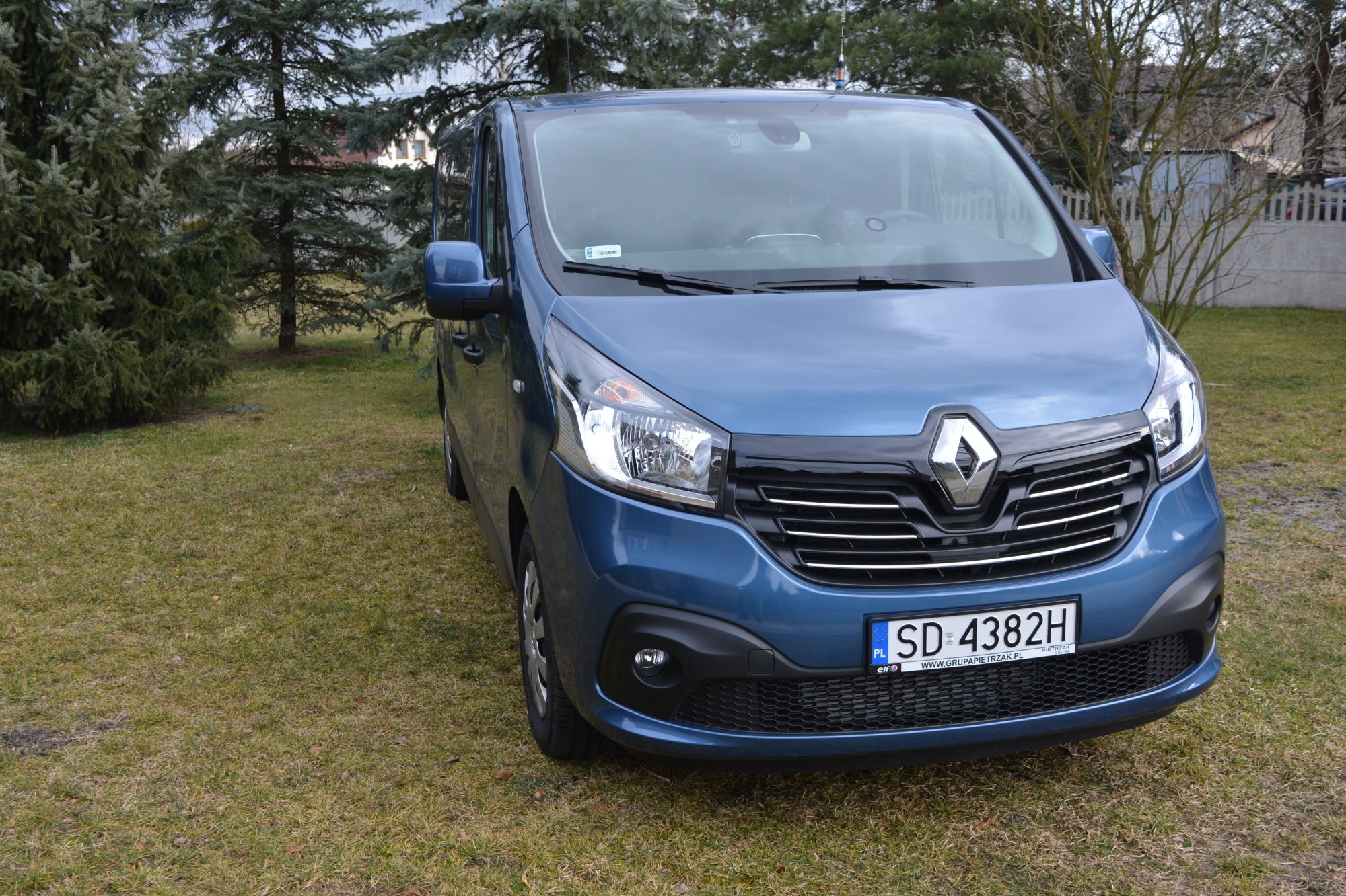 Niebieski van Renault z przodu, zaparkowany na trawniku przed drzewami, z widoczną tablicą rejestracyjną SD 4382H.