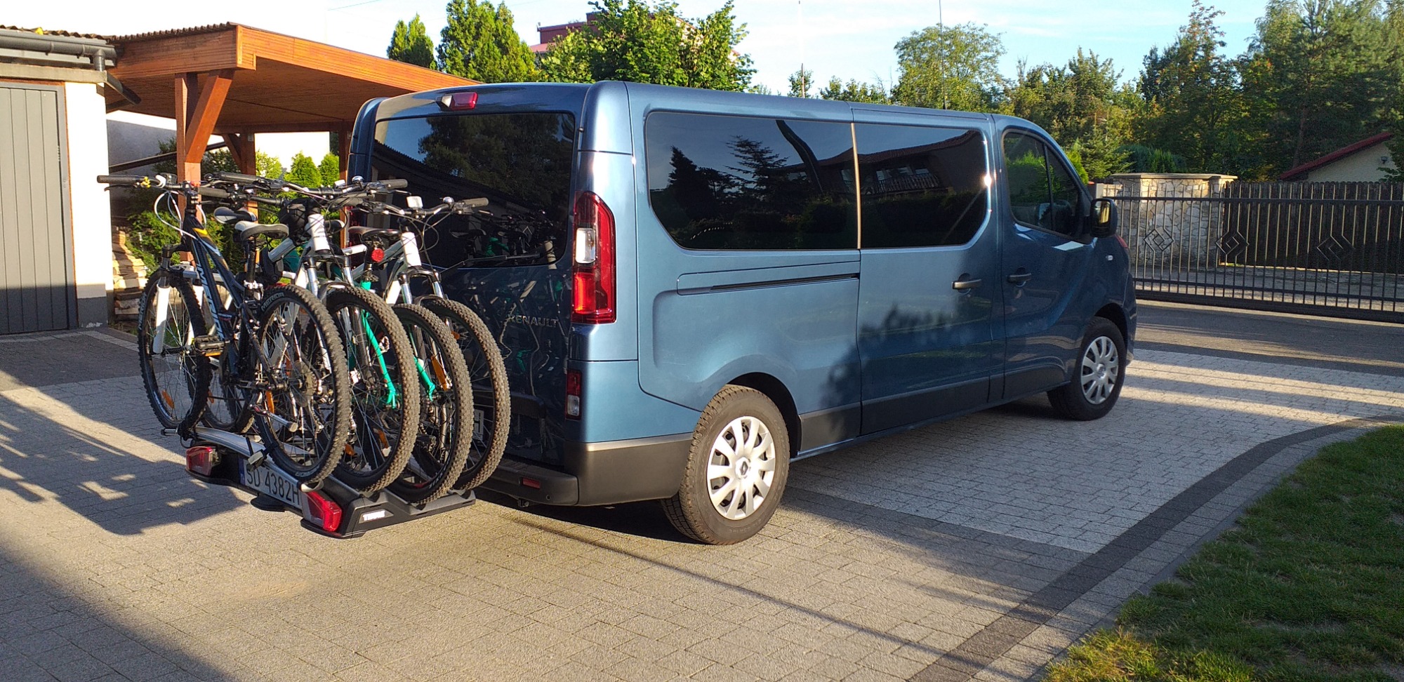 Niebieski van Renault z zamontowanym bagażnikiem rowerowym Thule, przewożący cztery rowery górskie na tle brukowanej posesji z zadaszeniem i ogrodzeniem.