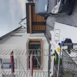 Dwukondygnacyjny dom z białej cegły, w trakcie przygotowań do prac elewacyjnych, z widocznym rusztowaniem, zabezpieczonym oknem i przygotowanymi płytami styropianowymi na podjeździe, obok...