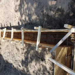 Szczegółowy widok deskowania fundamentu zbrojonego stalą, przygotowanego do zalania betonem, widoczne ślady stóp w piasku.