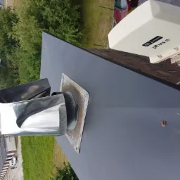 Nowa, grafitowa blacha na dachu z kominem i anteną satelitarną Plus, widok z góry na tle zieleni.