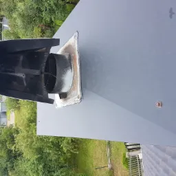 Szary, metalowy komin wentylacyjny z osłoną w kształcie daszka, zamontowany na płaskim dachu, widok z góry, z widocznym otoczeniem zieleni.