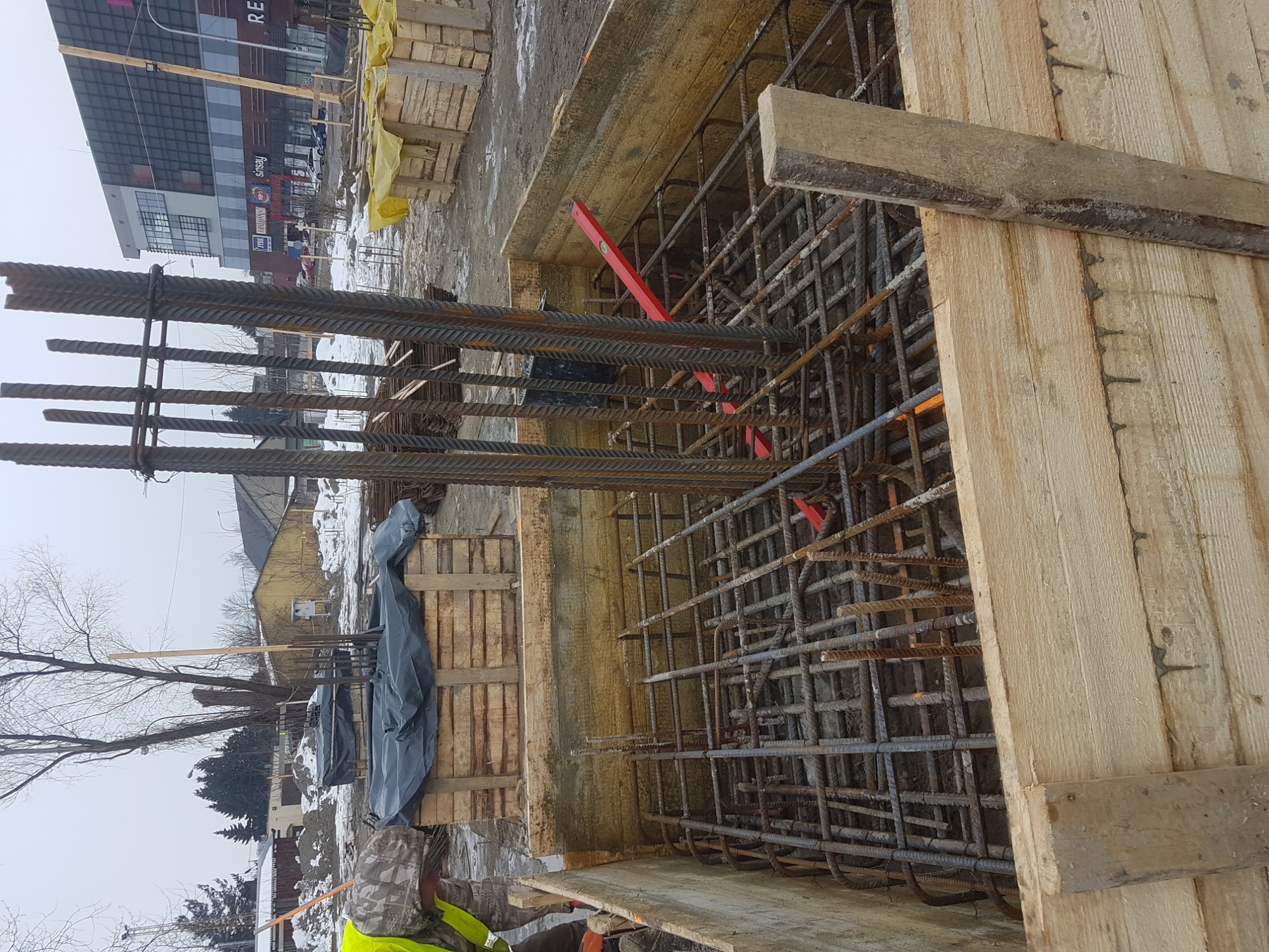 Zbrojenie fundamentów w drewnianym szalunku, widoczny poziomica i fragment budynku w tle.