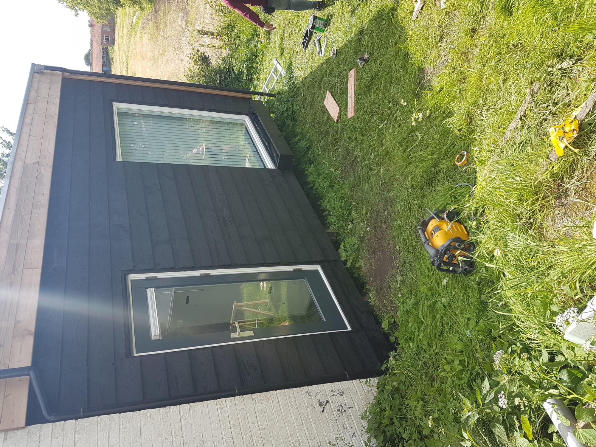 Czarny, drewniany budynek gospodarczy z oknem i drzwiami, widok z zewnątrz, na trawie leżą narzędzia, w tle zielona trawa i budynki.