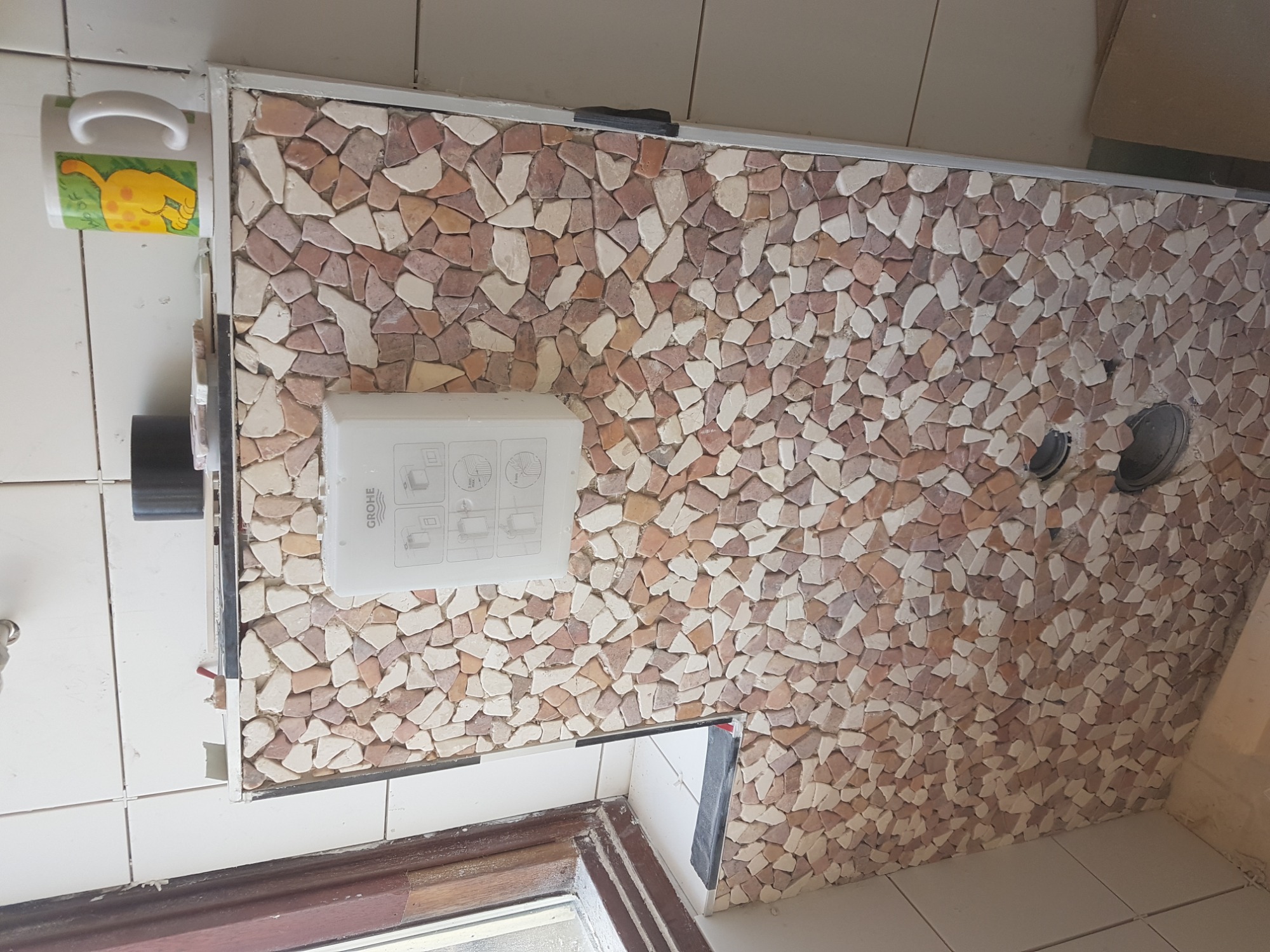 Ściana w łazience wykończona mozaiką z kamieni w odcieniach beżu i brązu, z widocznym przyciskiem spłuczki marki Grohe i kubkiem z motywem zwierzęcym zawieszonym na ścianie.