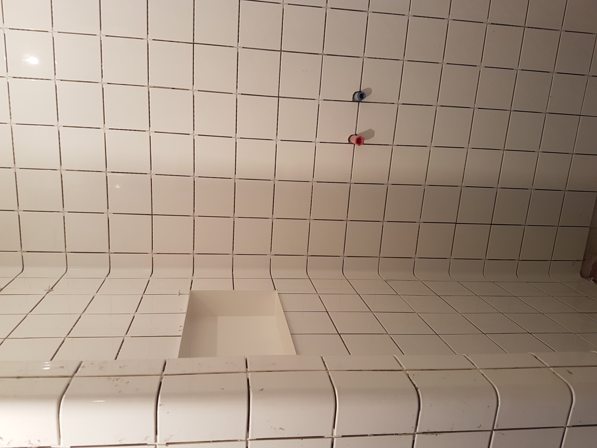 Ściany i półka w trakcie układania białych, kwadratowych płytek w łazience, widoczne wystające rurki z zaworami.