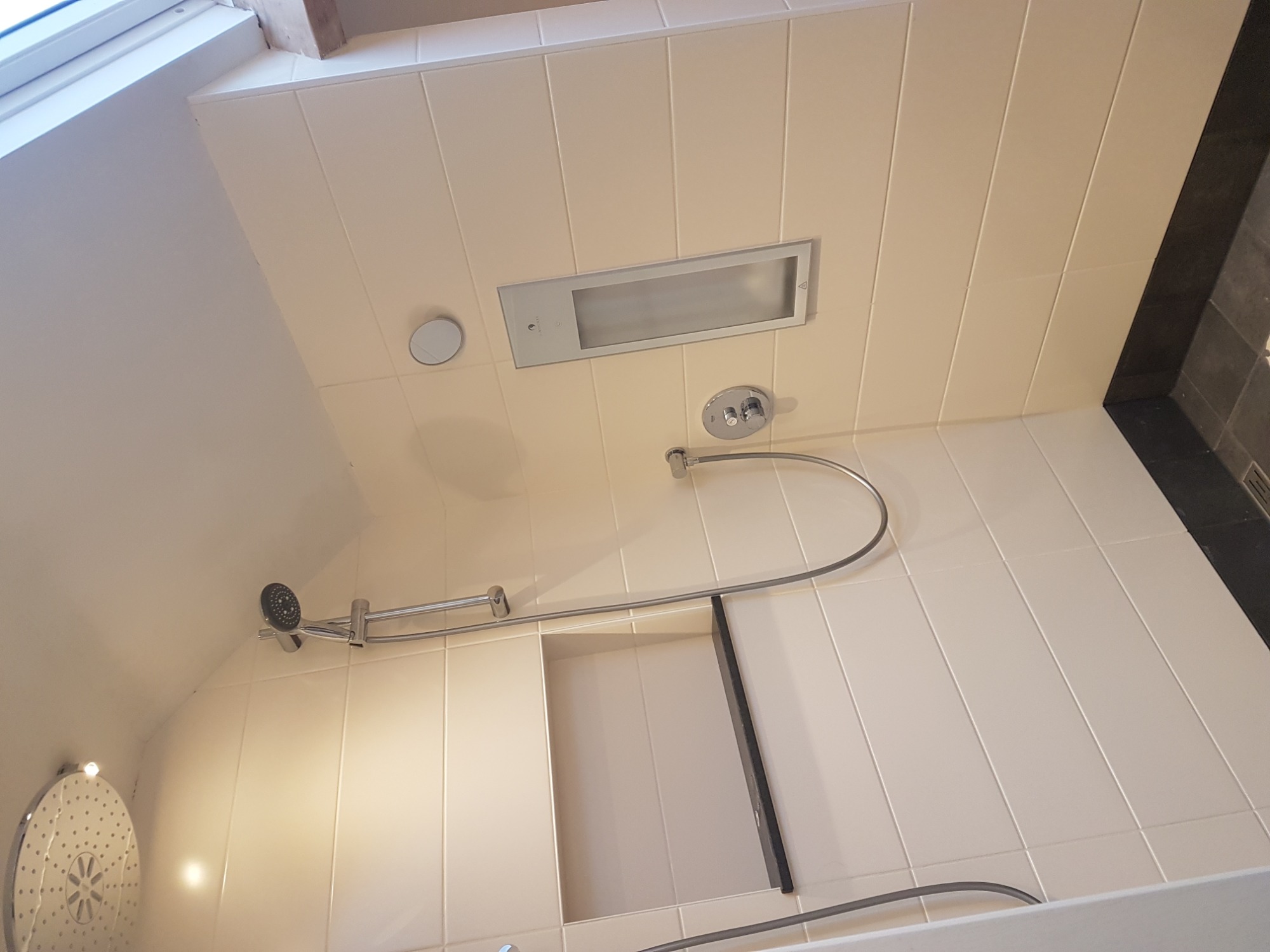 Wykończona łazienka z beżowymi płytkami, widoczna armatura prysznicowa, wnęka w ścianie z czarną obramówką, oświetlenie sufitowe i panel sterowania oświetleniem.