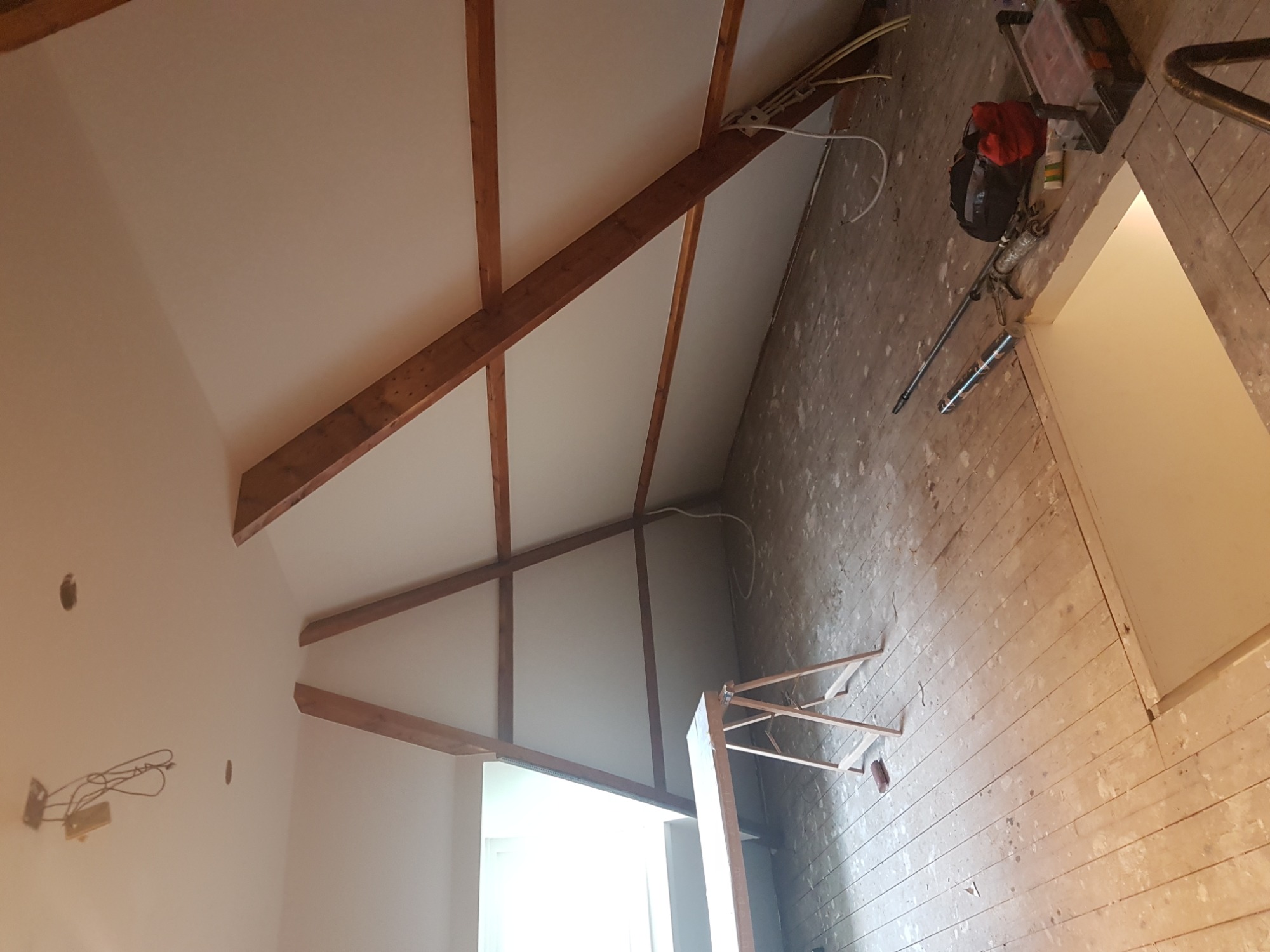 Poddasze w trakcie prac: widoczne drewniane belki konstrukcyjne, białe płyty kartonowo-gipsowe na skośnym suficie, odsłonięta instalacja elektryczna i narzędzia na podłodze z desek.