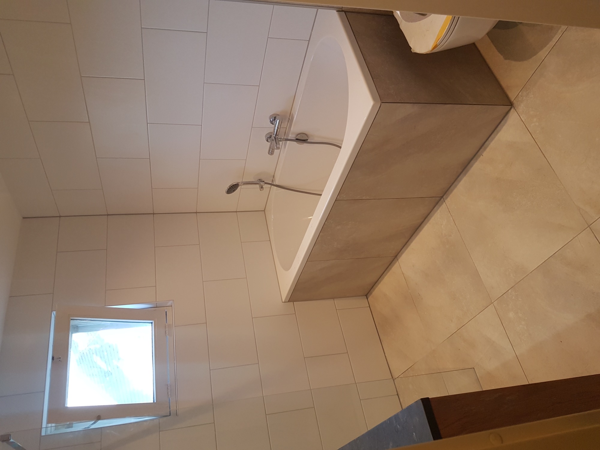 Wykończona łazienka z wanną obudowaną szarą płytką, białe płytki na ścianach, małe okno z otwartym skrzydłem, beżowa podłoga z płytek.