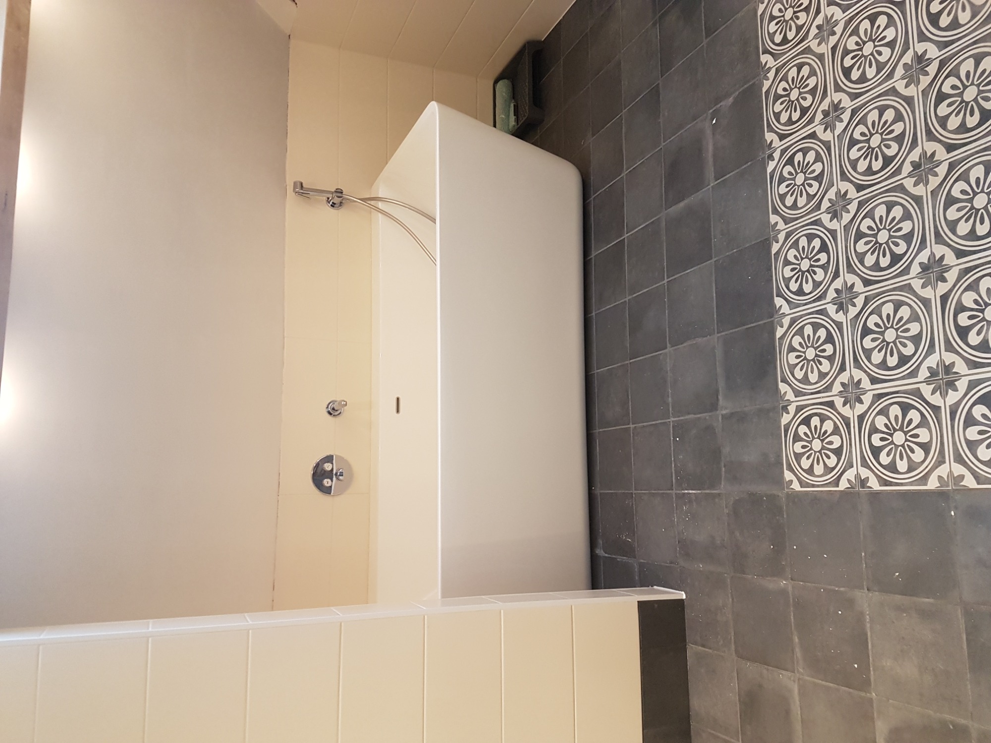 Nowoczesna łazienka z obudowaną wanną, kremowymi kafelkami na wannie i ścianie oraz ciemnymi, kwadratowymi płytkami z dekoracyjnym motywem kwiatowym na ścianie.