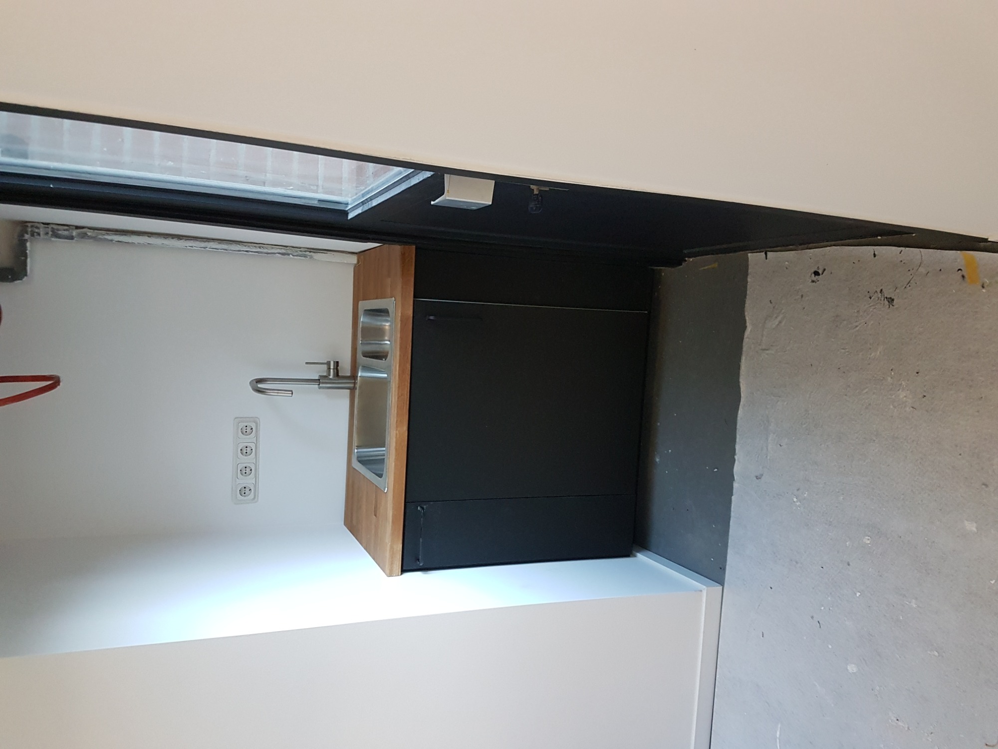 Narożny, podwójny zlew ze stali nierdzewnej z drewnianym blatem, wbudowany w czarną szafkę, zamontowany na białej ścianie z czterema gniazdkami elektrycznymi, widoczny fragment surowej ściany.