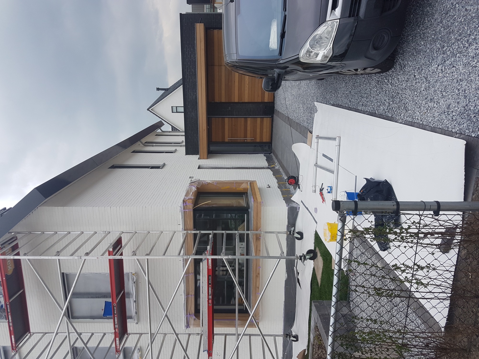 Dwukondygnacyjny dom z białej cegły, w trakcie przygotowań do prac elewacyjnych, z widocznym rusztowaniem, zabezpieczonym oknem i przygotowanymi płytami styropianowymi na podjeździe, obok...