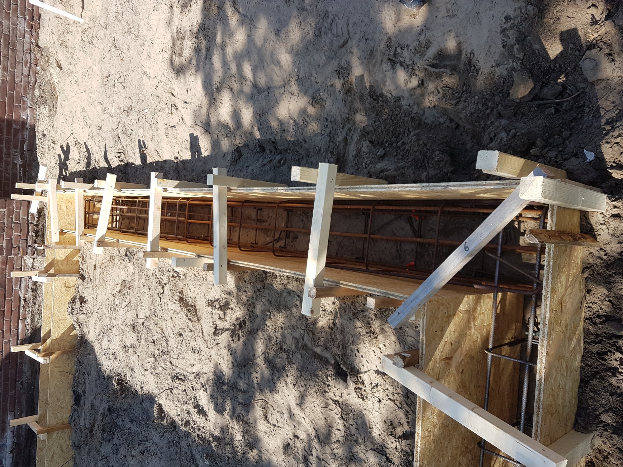 Szczegółowy widok deskowania fundamentu zbrojonego stalą, przygotowanego do zalania betonem, widoczne ślady stóp w piasku.