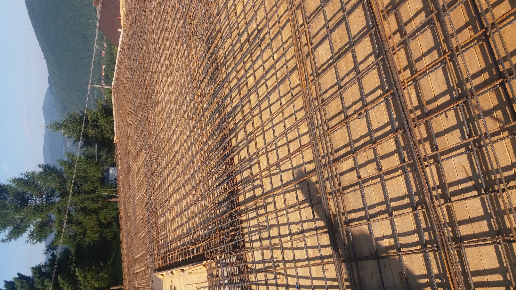 Zbrojenie płyty fundamentowej z widoczną siatką stalową i drewnianym szalunkiem, w tle zalesione wzgórza.