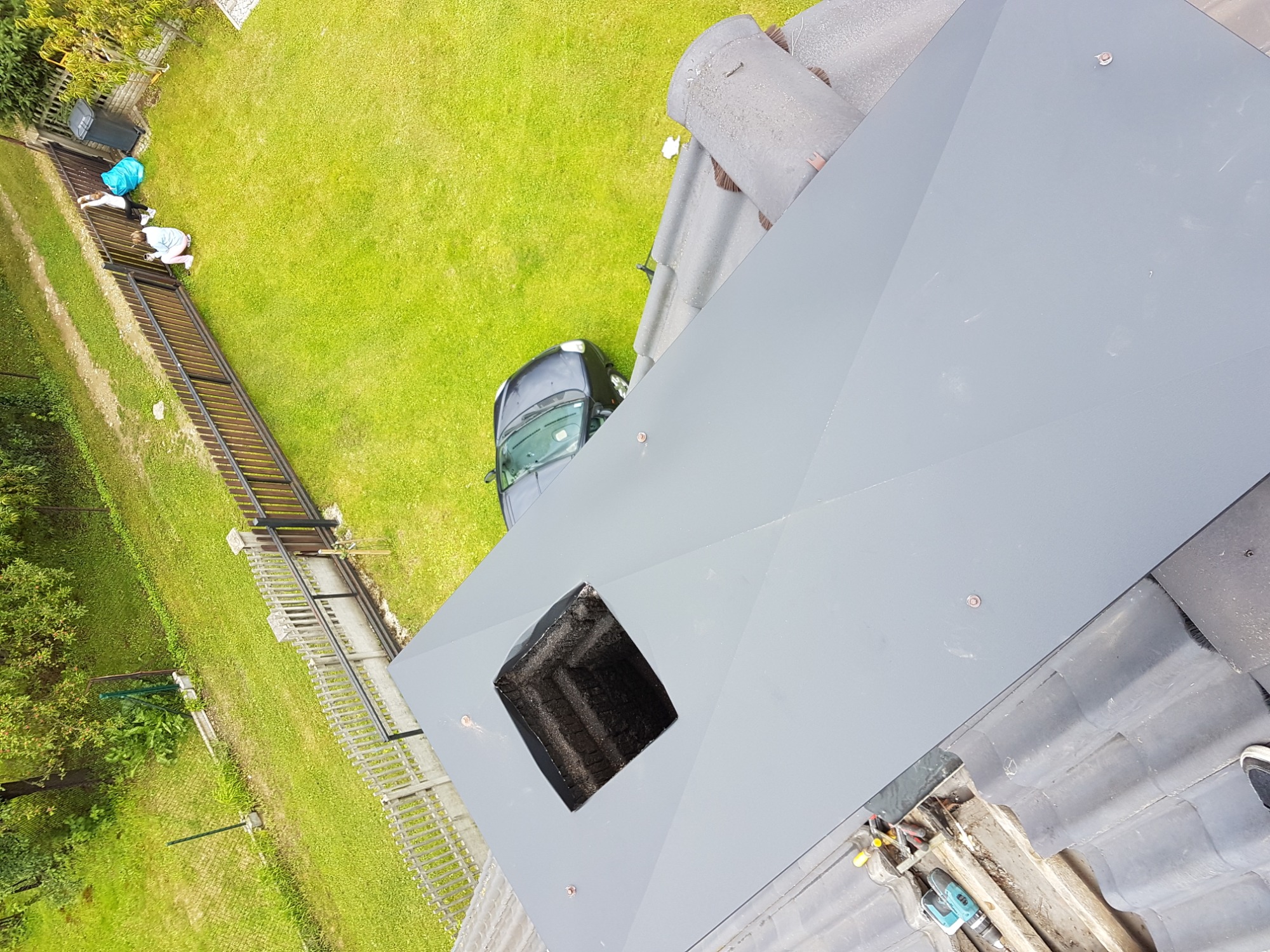 Widok z góry na nową, szarą obudowę komina z otworem, narzędzia i fragment dachu z dachówką, trawnik, samochód i osoby przy płocie w tle.