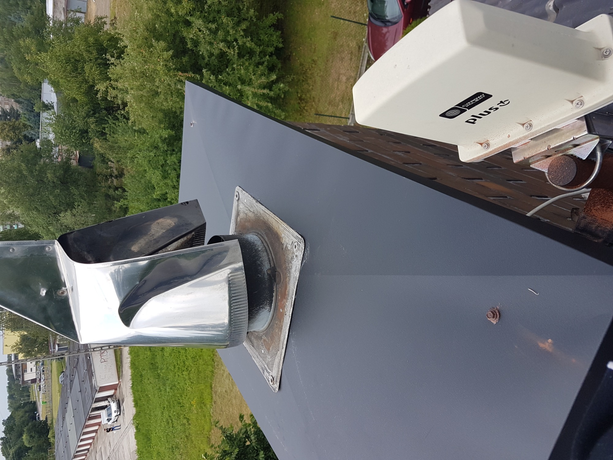 Nowa, grafitowa blacha na dachu z kominem i anteną satelitarną Plus, widok z góry na tle zieleni.
