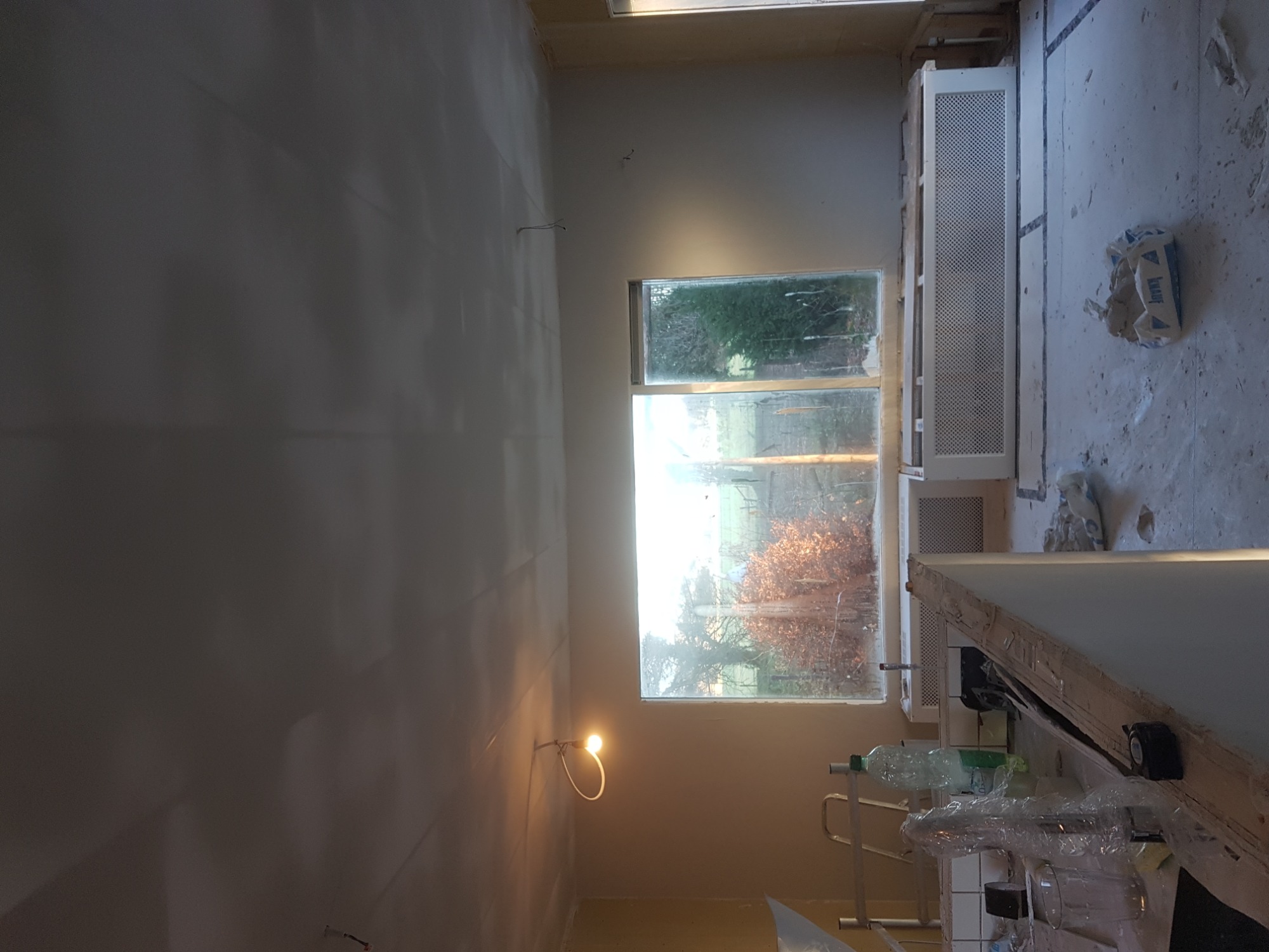 Wnętrze w trakcie prac wykończeniowych: ściany pokryte gładzią, widoczne okno z widokiem na zewnątrz, prowizoryczne oświetlenie, resztki materiałów budowlanych i narzędzia.
