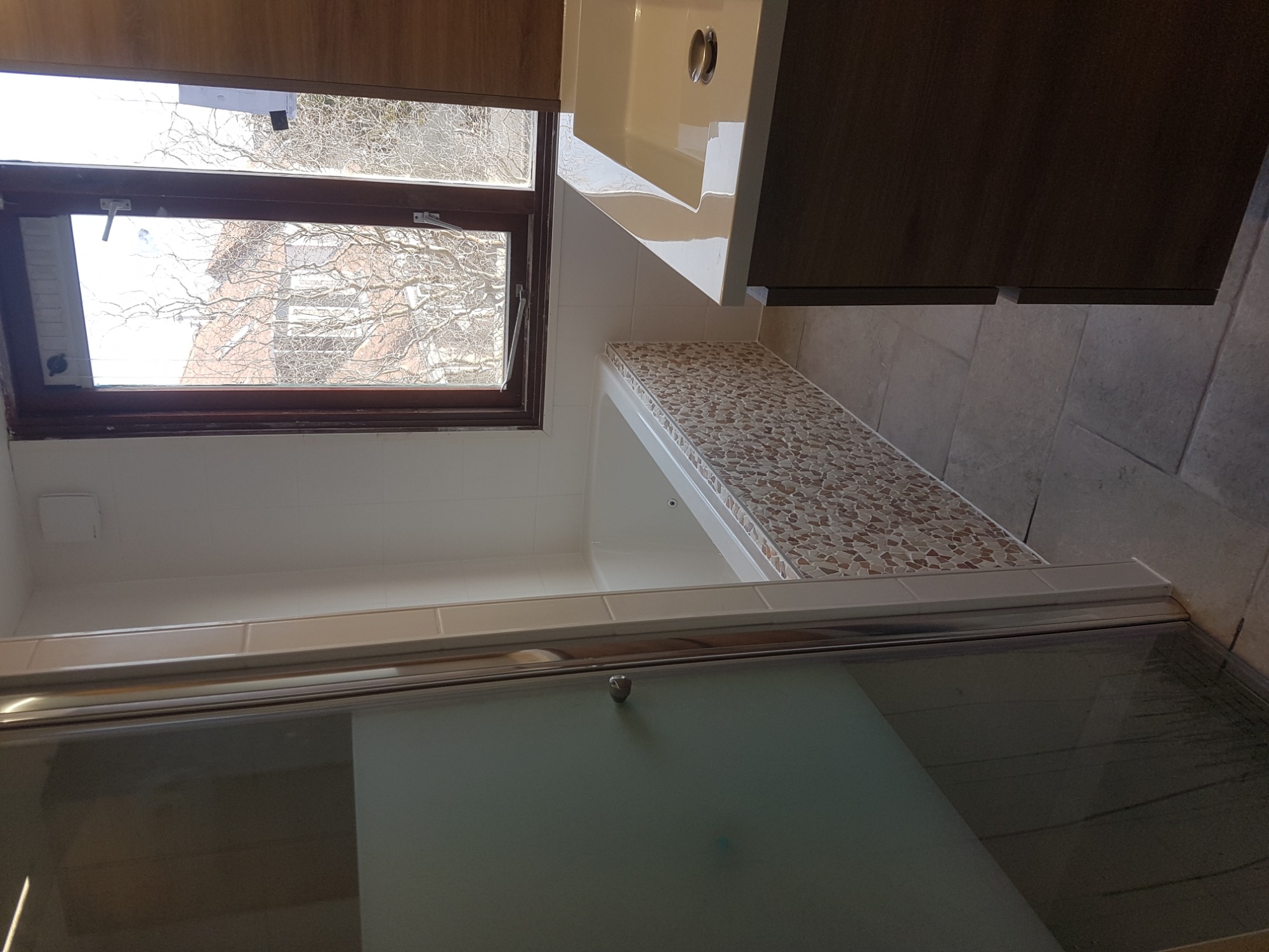 Nowoczesna łazienka po remoncie: kabina prysznicowa z przesuwnymi drzwiami, mozaika na obudowie wanny, okno z widokiem na drzewa, umywalka wbudowana w szafkę z drewnianymi frontami, szare płytki...