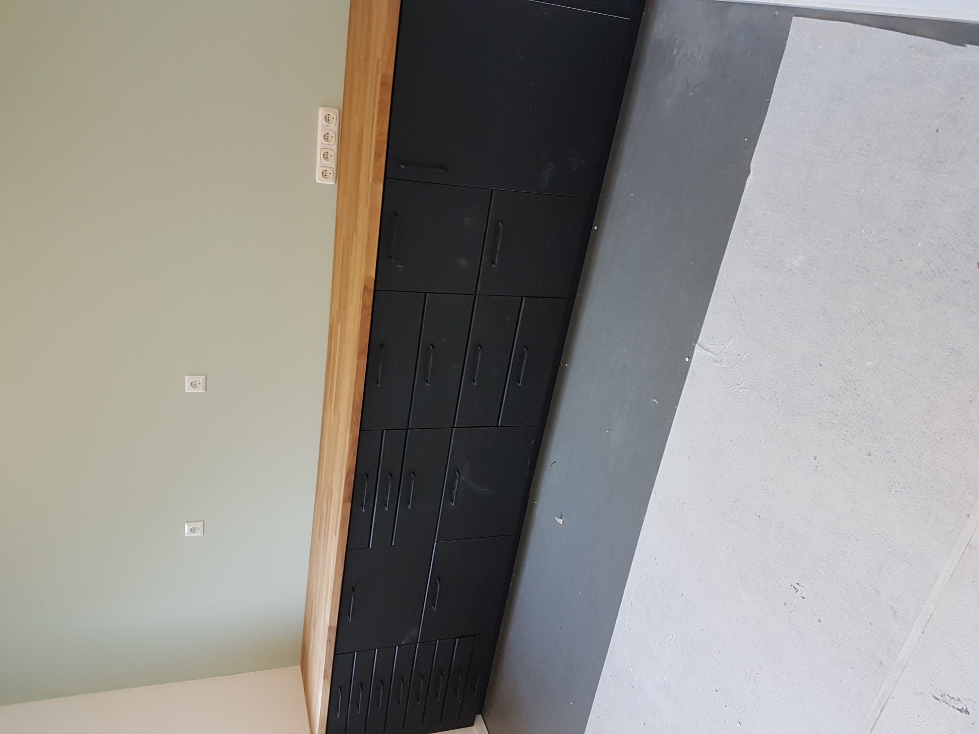 Ciemne szafki z drewnianym blatem w jasnym pomieszczeniu z szarą podłogą, widoczne gniazdka elektryczne na ścianie.