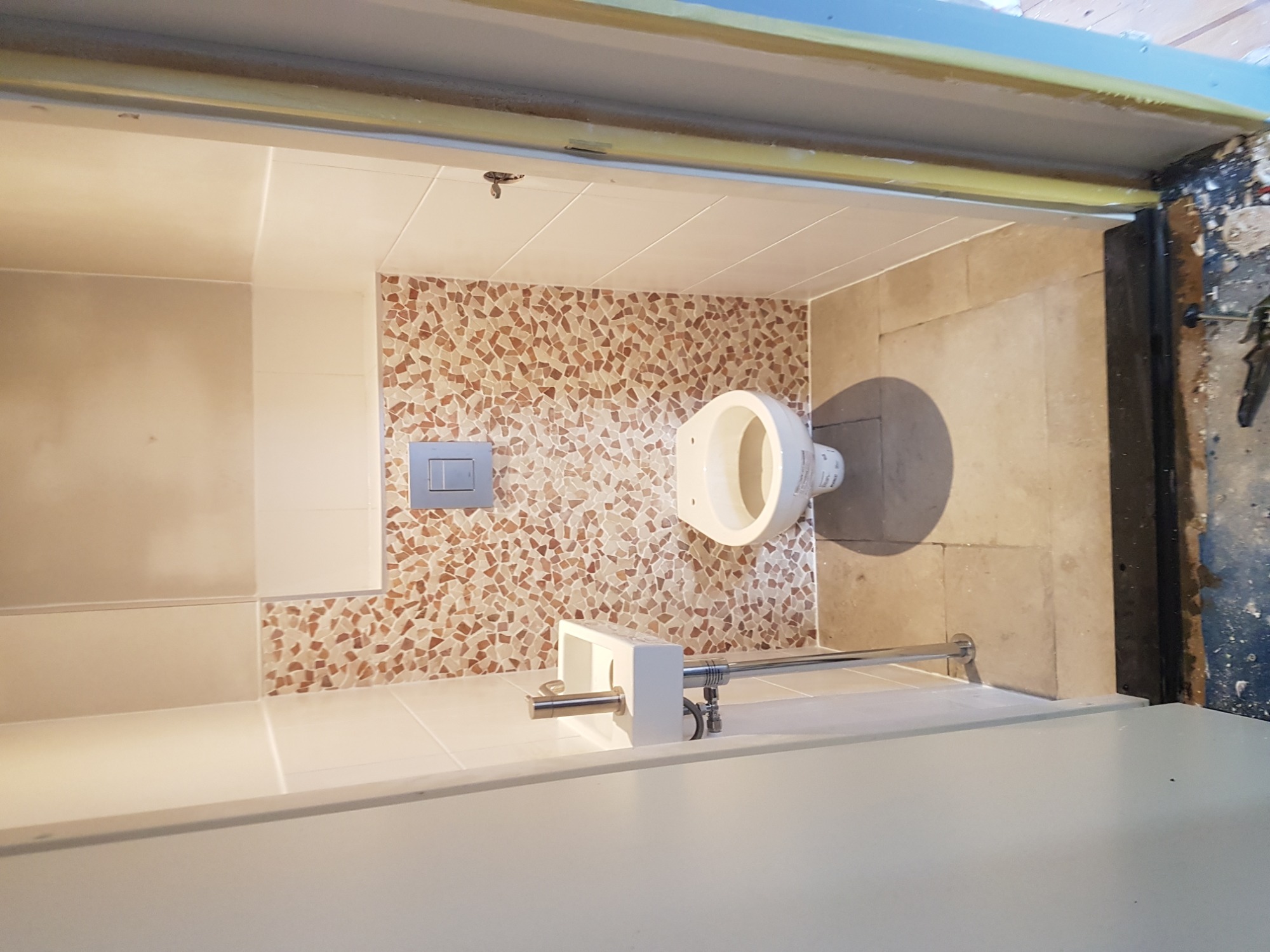 Wnętrze łazienki w trakcie prac wykończeniowych, widoczna mozaika na ścianie, biała ceramika sanitarna i fragment instalacji wodnej, surowy stan wykończenia przy framudze drzwi.