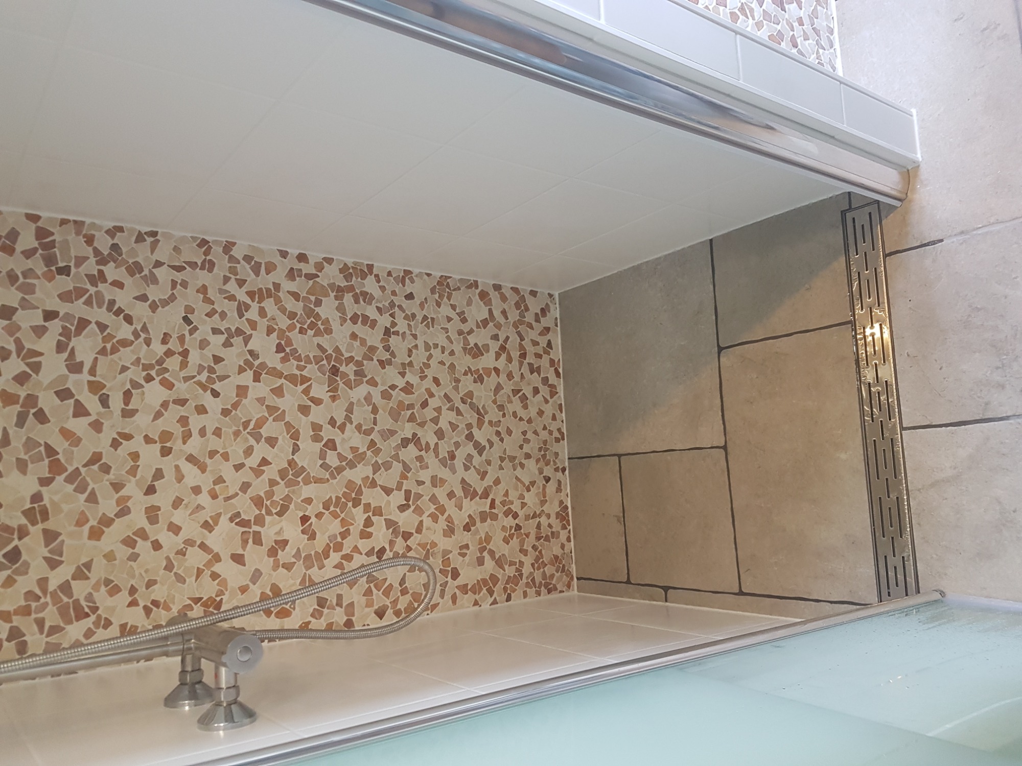 Fragment wykończonej łazienki z mozaikową ścianą, baterią prysznicową i szklanym panelem kabiny prysznicowej.