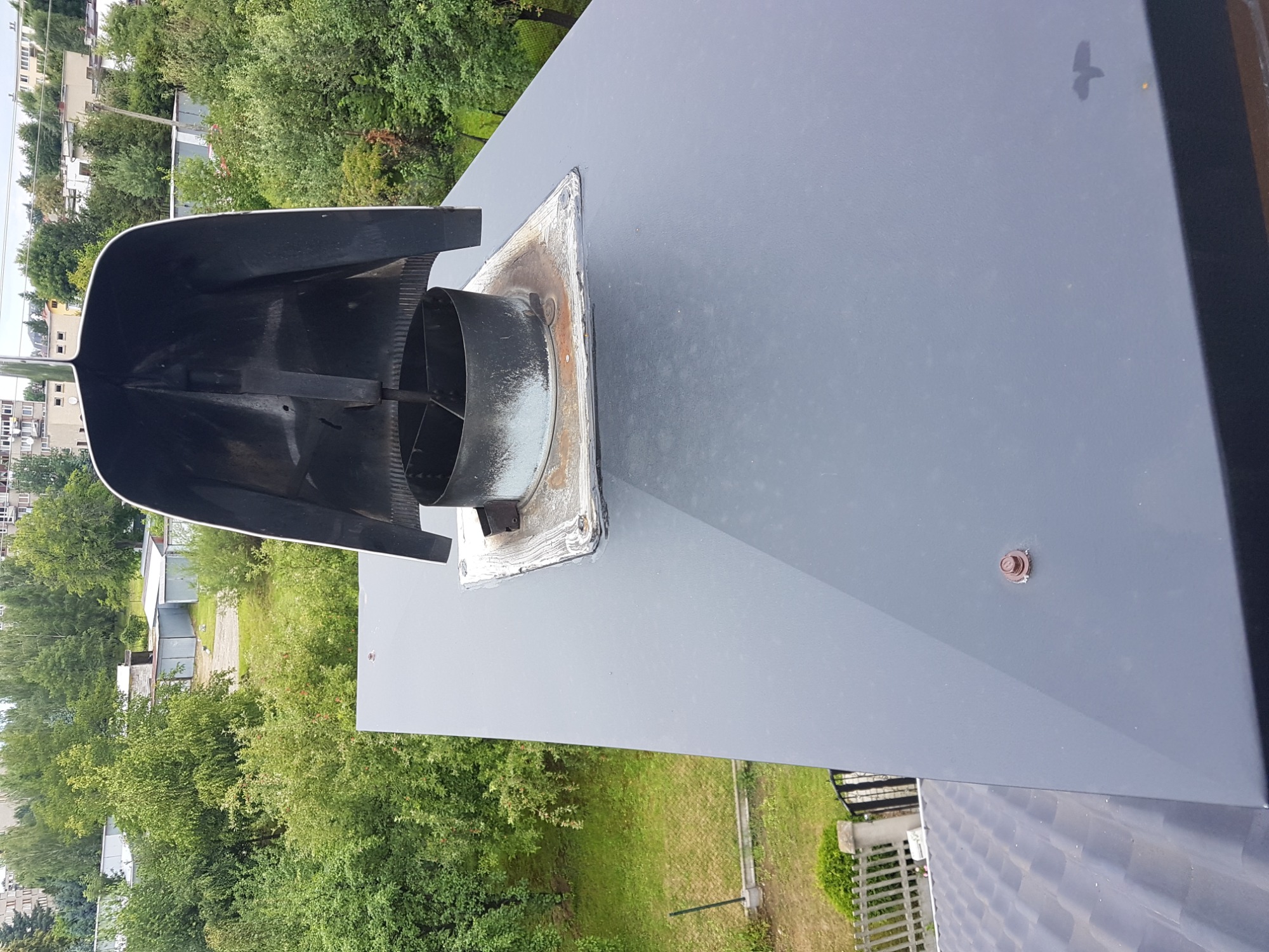 Szary, metalowy komin wentylacyjny z osłoną w kształcie daszka, zamontowany na płaskim dachu, widok z góry, z widocznym otoczeniem zieleni.