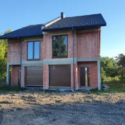 Surowy stan dwukondygnacyjnego domu z czerwonej cegły z brązowymi bramami garażowymi i drzwiami wejściowymi, czarny dach, teren wokół domu nierówny, w tle zieleń drzew.