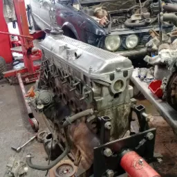 Wyjęty silnik samochodowy na czerwonym stojaku, obok częściowo rozebrany samochód marki BMW w warsztacie samochodowym.