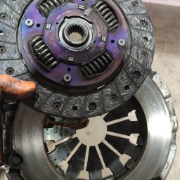 Rama Garage - Zużyte sprzęgło samochodowe z widocznymi śladami przegrzania (fioletowe zabarwienie) i śladami użytkowania, trzymane w rękawicy roboczej.