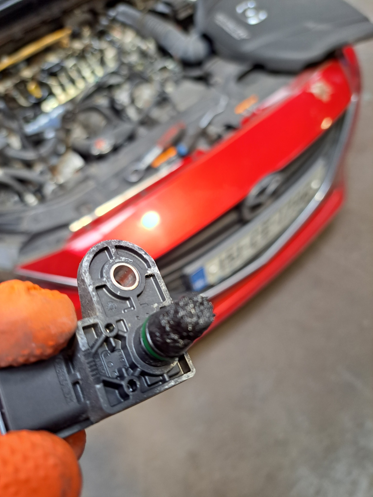 Czujnik zanieczyszczony nagarem trzymany w pomarańczowej rękawicy, na tle otwartej maski czerwonej Mazdy z silnikiem diesla 2.2 Skyactiv, sugerujący problem z ciśnieniem oleju i utratą mocy.