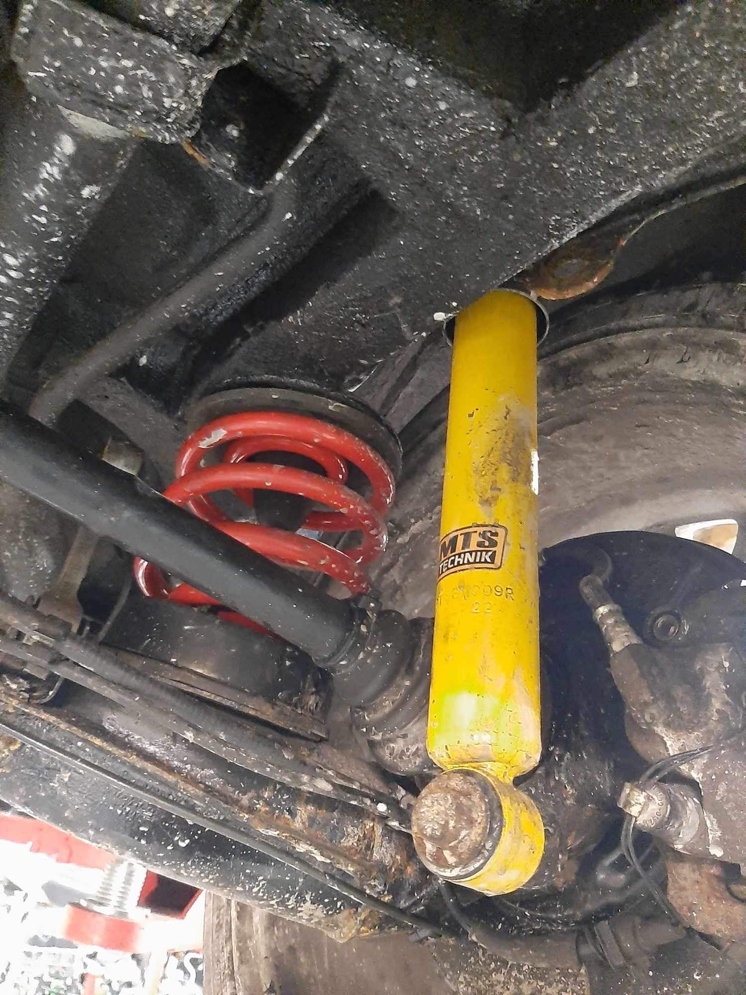 Podwozie samochodu z widocznym żółtym amortyzatorem MTS Technik i czerwoną sprężyną zawieszenia, sfotografowane z dołu, z widocznymi śladami eksploatacji i zabrudzeniami.