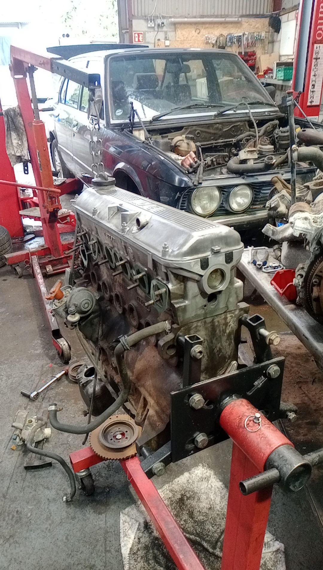 Wyjęty silnik samochodowy na czerwonym stojaku, obok częściowo rozebrany samochód marki BMW w warsztacie samochodowym.