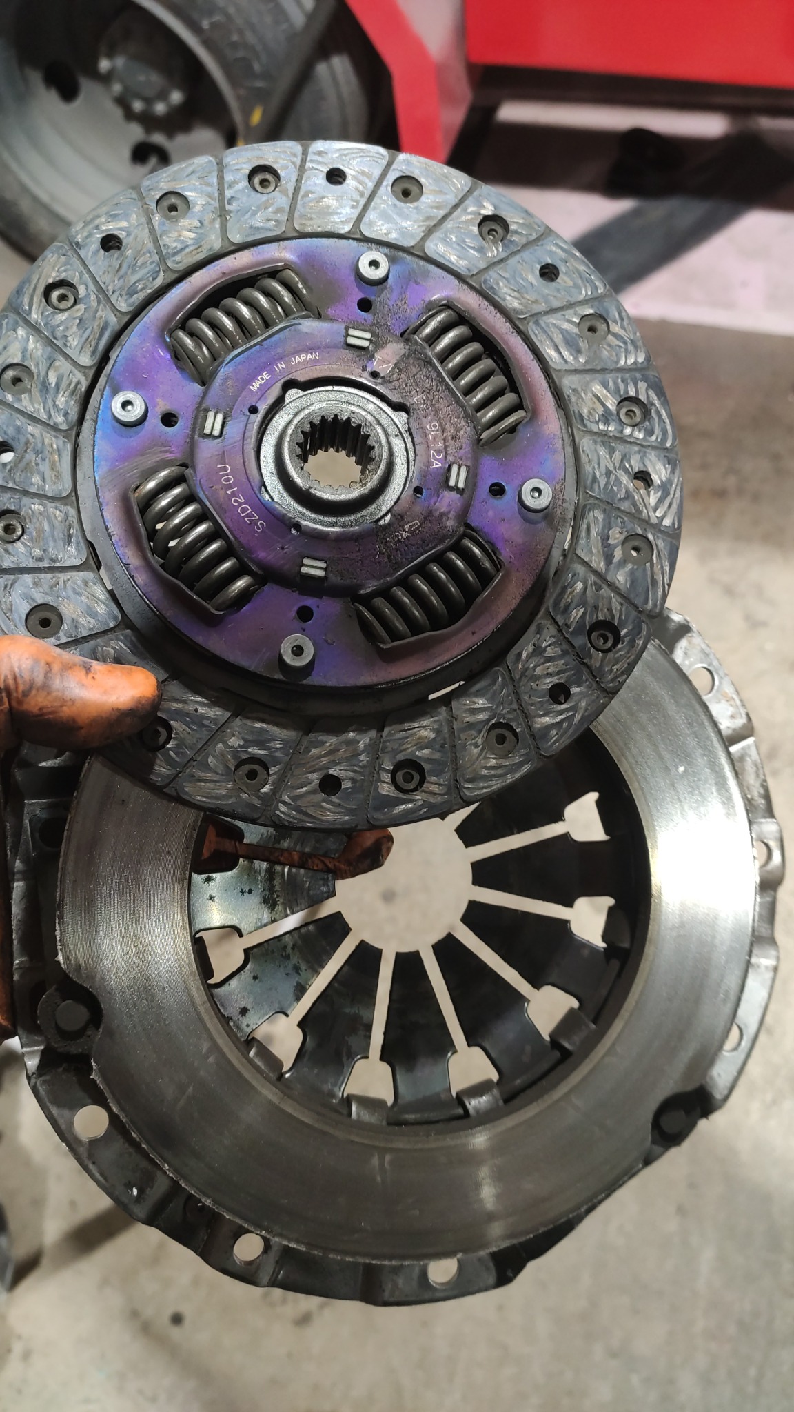 Zużyte sprzęgło samochodowe z widocznymi śladami przegrzania (fioletowe zabarwienie) i śladami użytkowania, trzymane w rękawicy roboczej.
