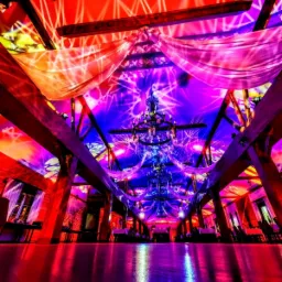 Uroczysta sala z drewnianą konstrukcją, oświetlona kolorowymi światłami i ozdobiona zwisającymi tkaninami, z parkietem odbijającym światło.