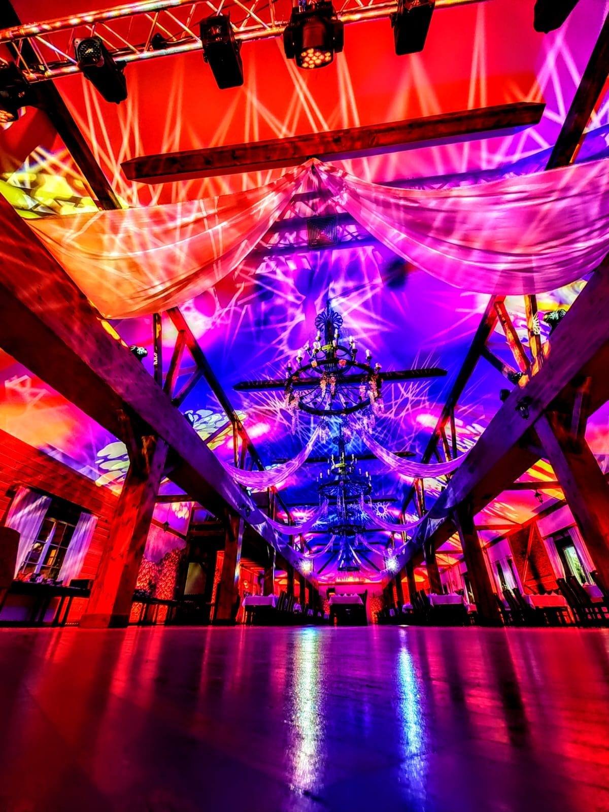 Uroczysta sala z drewnianą konstrukcją, oświetlona kolorowymi światłami i ozdobiona zwisającymi tkaninami, z parkietem odbijającym światło.