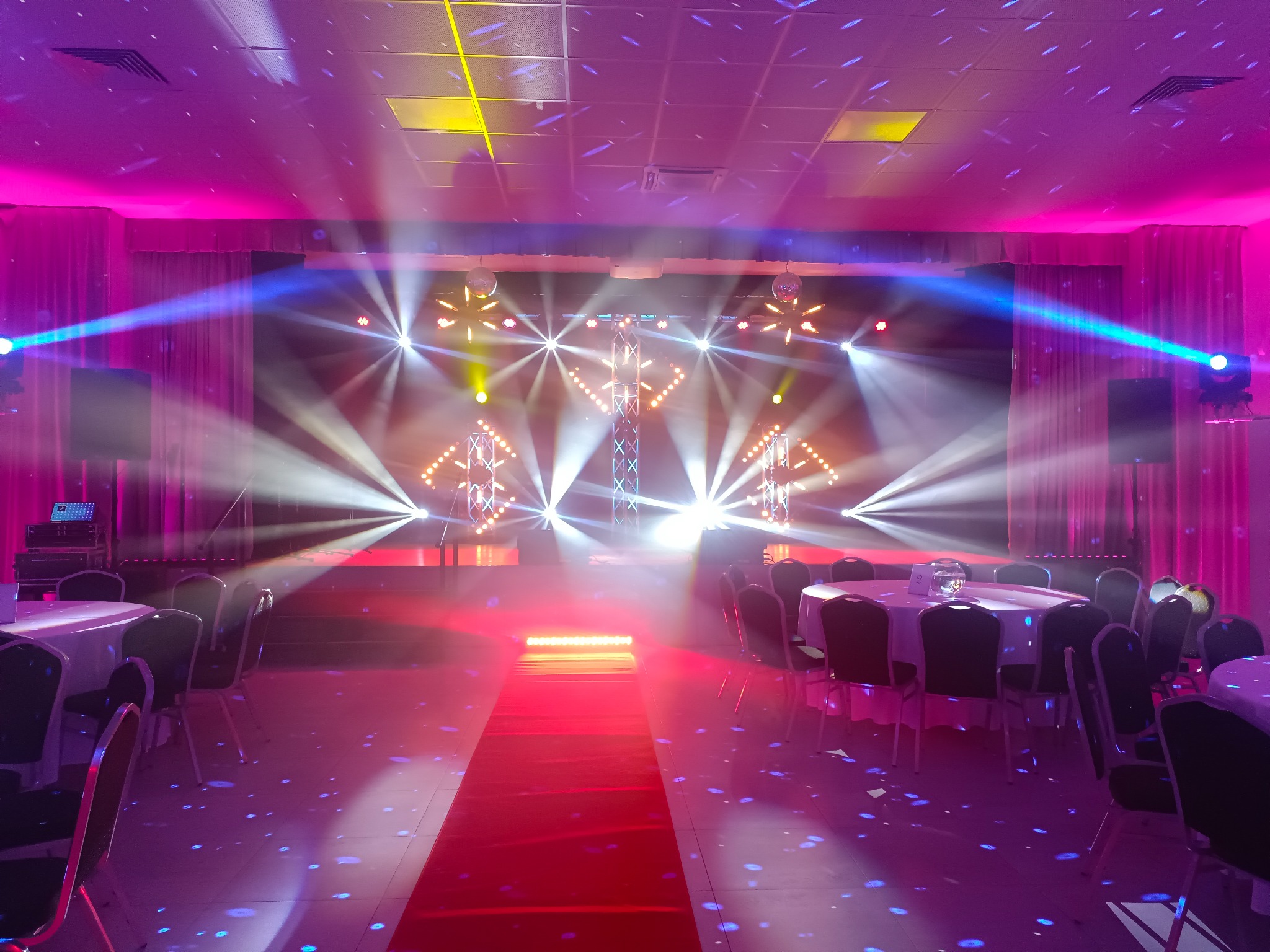 Sala bankietowa z dekoracją świetlną: scena z reflektorami, czerwony dywan, okrągłe stoły z białymi obrusami, światła dyskotekowe na podłodze, różowe kurtyny.