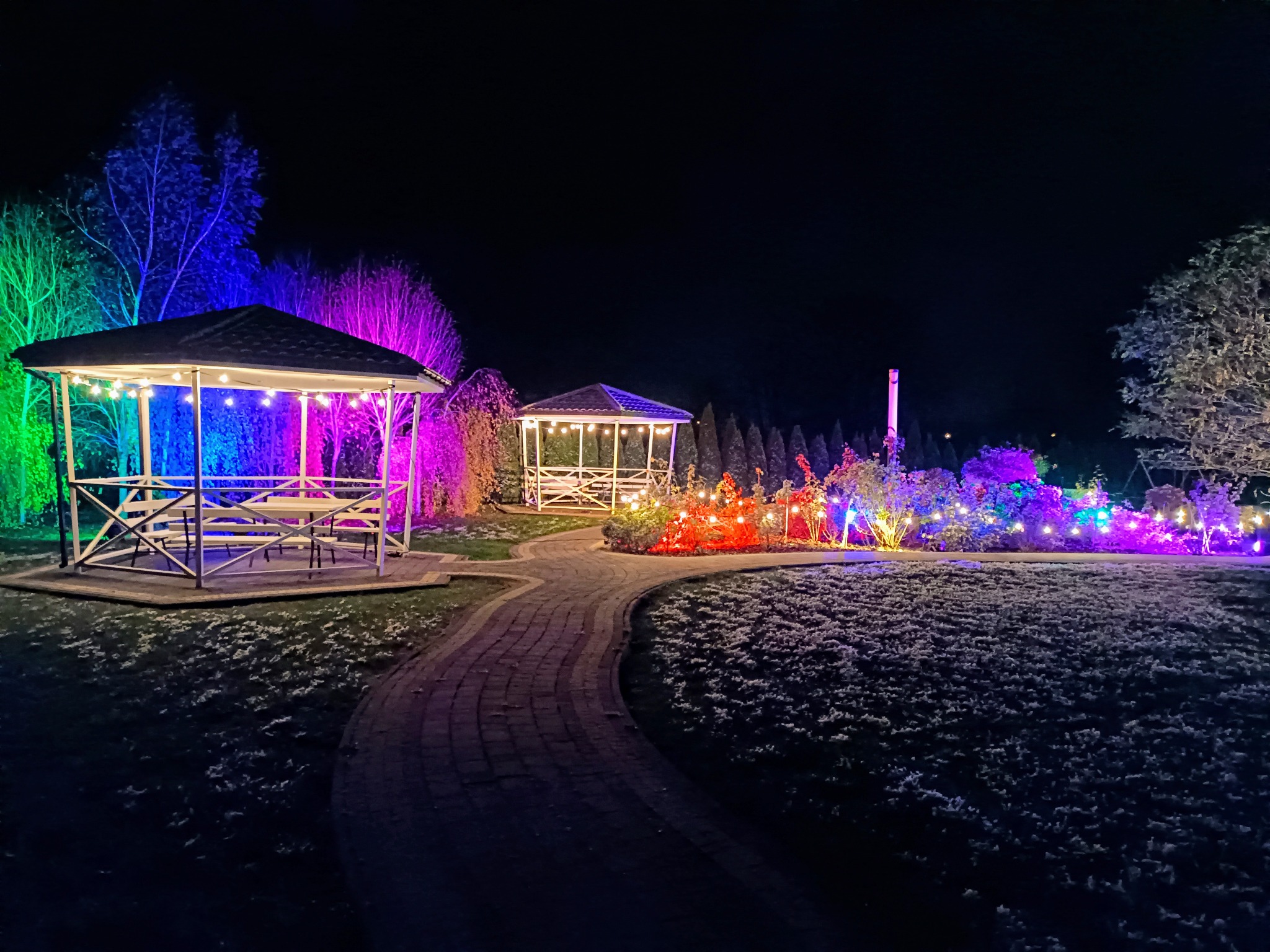 Dwie altany ogrodowe oświetlone różnokolorowymi światłami tworzą magiczną atmosferę nocą. Ścieżka prowadzi przez ogród z dekoracyjnym oświetleniem.