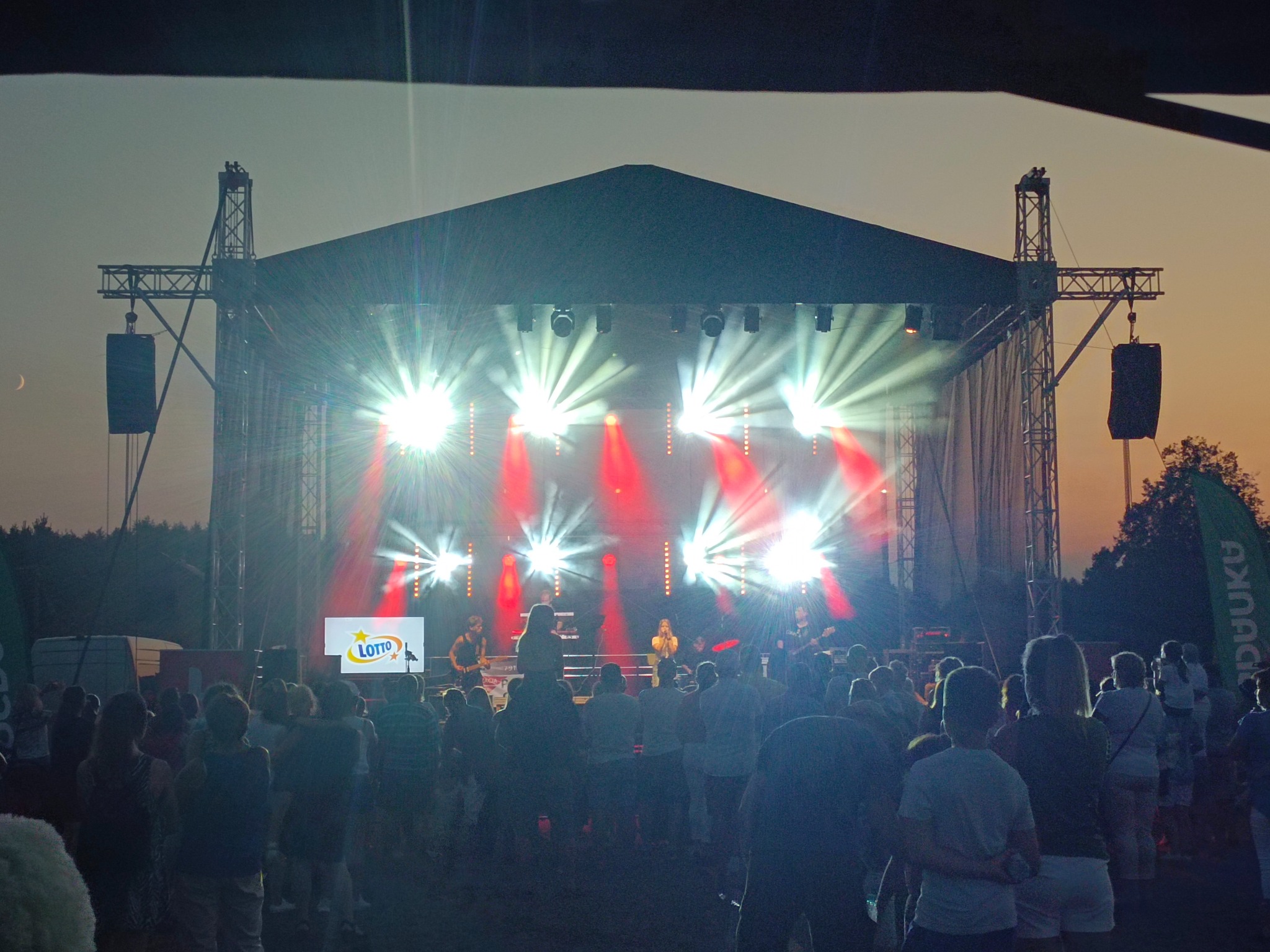 Scena plenerowa z zespołem muzycznym i publicznością, intensywne światła reflektorów, logo Lotto na ekranie. Wieczorny koncert na świeżym powietrzu.