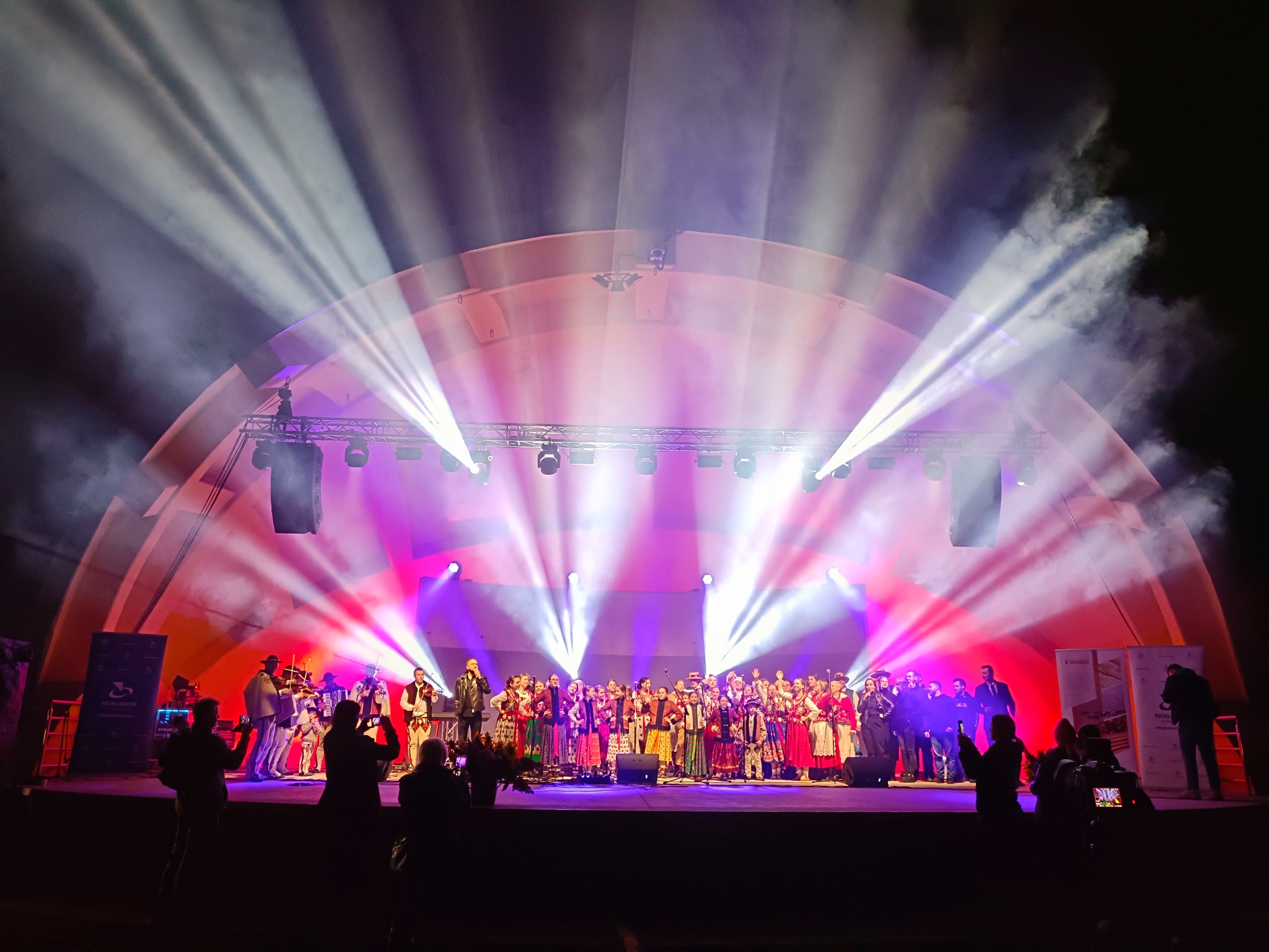 Scena z koncertem w Starym Majdanie, oświetlona reflektorami i światłami w kolorach różu i bieli. Na scenie zespół ludowy w strojach regionalnych.