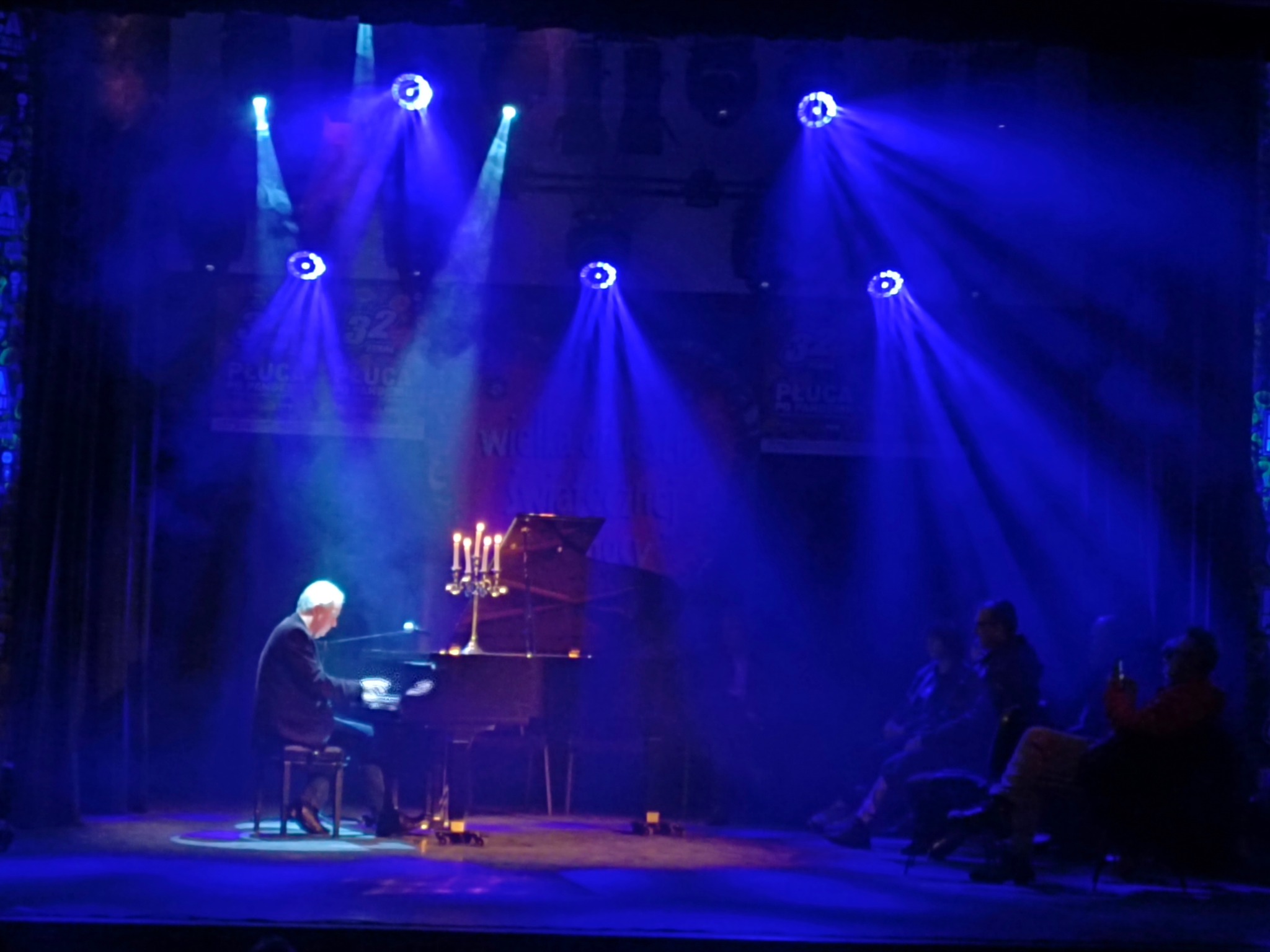 Scena z pianistą grającym na fortepianie, oświetlona niebieskimi reflektorami i świecami. W tle widoczna publiczność.