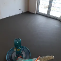 Świeżo wylana, gładka posadzka w jasnym pomieszczeniu z dużym oknem, częściowo widoczna maszyna do zacierania betonu.