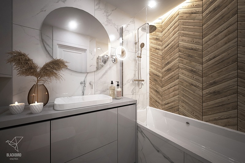 Nowoczesna łazienka z drewnianą ścianą ułożoną w jodełkę, okrągłe lustro nad prostokątną umywalką nablatową, marmurowe płytki na ścianach, dekoracyjne świece i trawa pampasowa w wazonie.