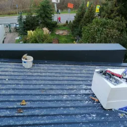 Styropian, pistolet do pianki, kielnia i wiadro na blaszanym dachu podczas prac ociepleniowych, widok z góry na otoczenie budynku.