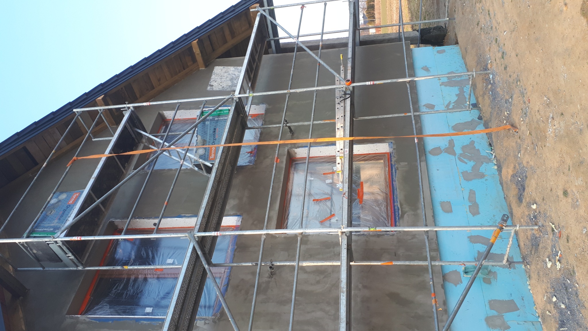 Dom w trakcie prac elewacyjnych: otynkowana ściana szczytowa z rusztowaniem, okna zabezpieczone folią, widoczne fragmenty ocieplenia fundamentów niebieskim styropianem.