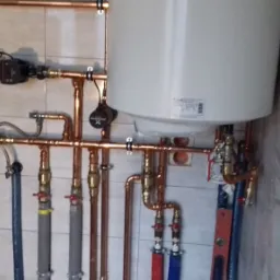 Nowoczesna instalacja grzewcza z miedzianymi rurami, białym zasobnikiem ciepłej wody, pompą obiegową i zaworami odcinającymi, zamontowana na ścianie wyłożonej płytkami ceramicznymi.